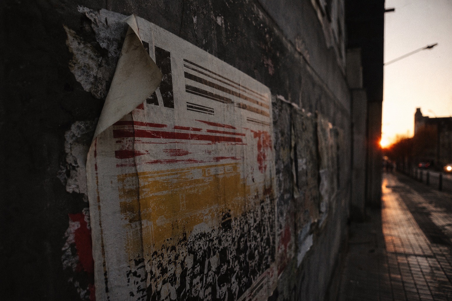 Un cartel de festival pegado en un muro urbano descascarillado, fotografiado en escorzo al atardecer, con una esquina despegada y restos de otros carteles debajo.