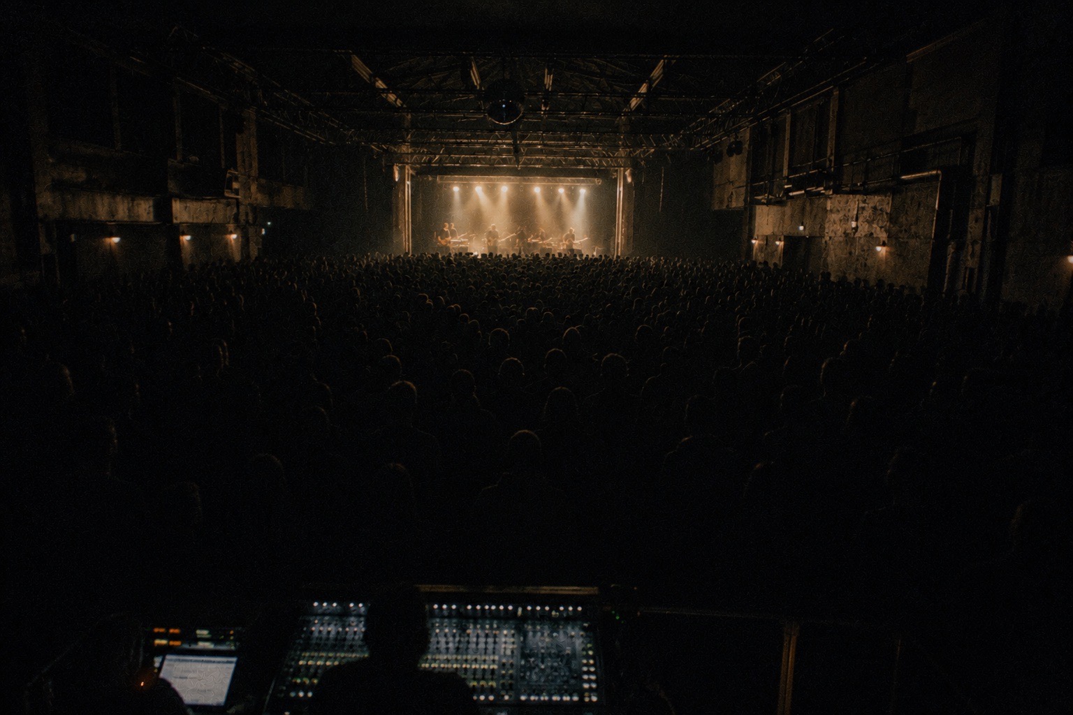 Una sala de conciertos llena vista desde la mesa de sonido: el público en penumbra, de espaldas, y el escenario iluminado al fondo.