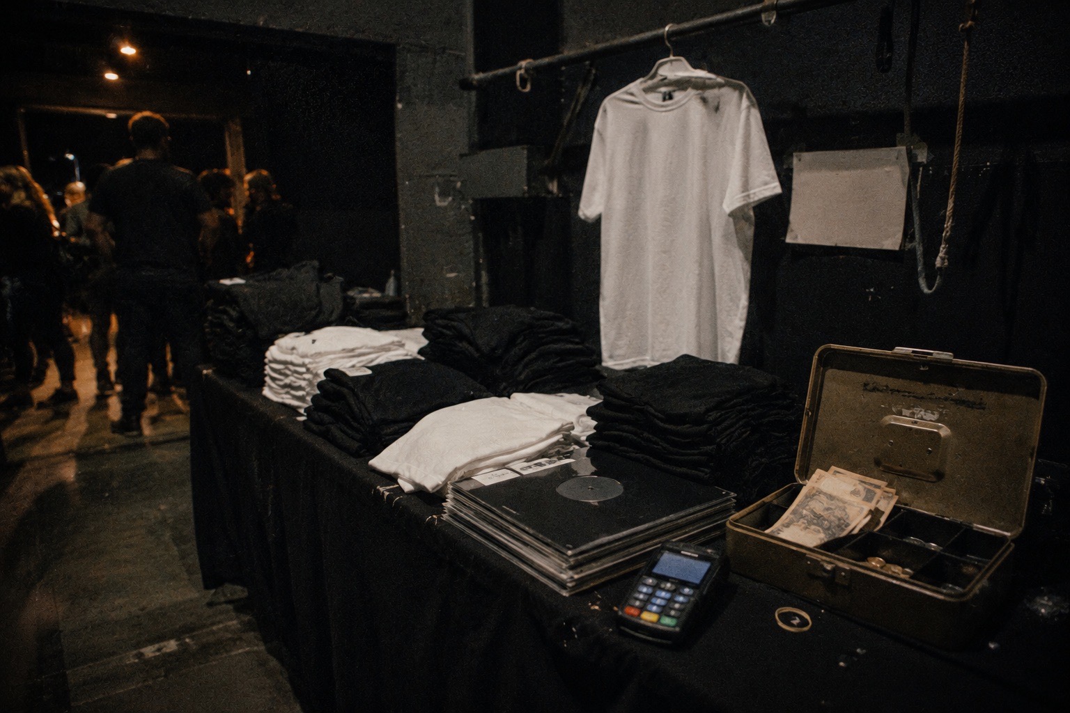 La mesa de merchandising al final de un concierto: camisetas dobladas en pilas, vinilos, un datafono y una caja metálica abierta con billetes, con la gente saliendo al fondo.